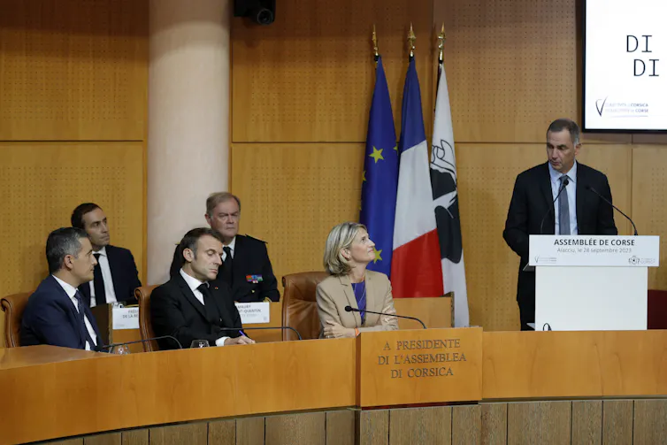 On voit un homme debout, le président du conseil exécutif de Corse Gilles Simeoni prononçant un discours devant le président Macron et le ministre français de l'Intérieur Gérald Darmanin qui l'écoutent.