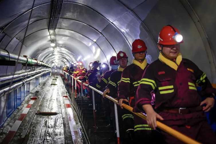 Workers in hard hats and overalls descend a mine shaft.