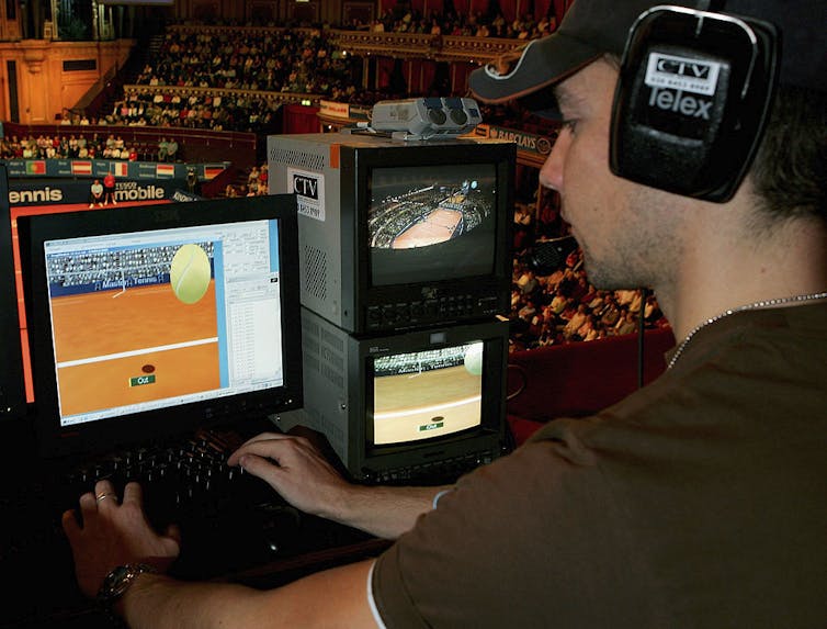 A man wearing a headset works on a computer with a graphic of a tennis ball and a court on the computer screen.