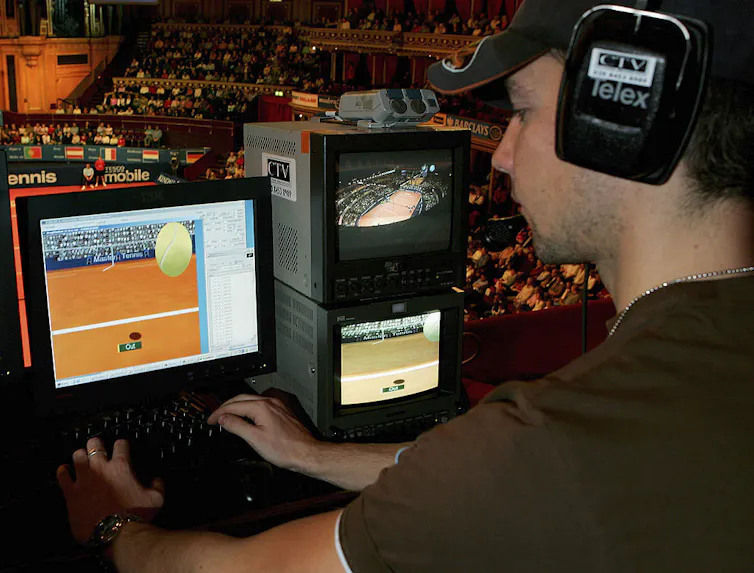 A man wearing a headset works on a computer with a graphic of a tennis ball and a court on the computer screen.