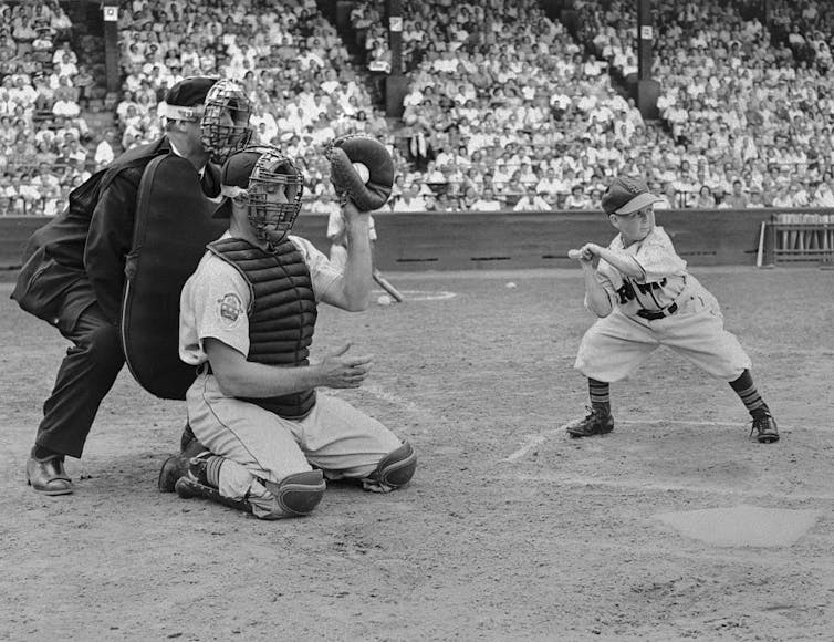 Black-and-white photo of very short man holding a bat in front of a kneeling catcher and crouching umpire.