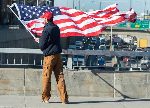 A man in a red ball cap is photographed from behind waving an American flag from an overpass.