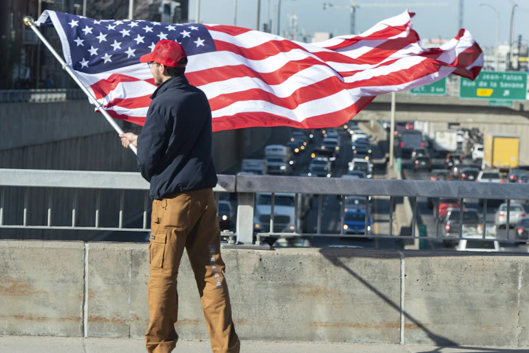 A man in a red ball cap is photographed from behind waving an American flag from an overpass.