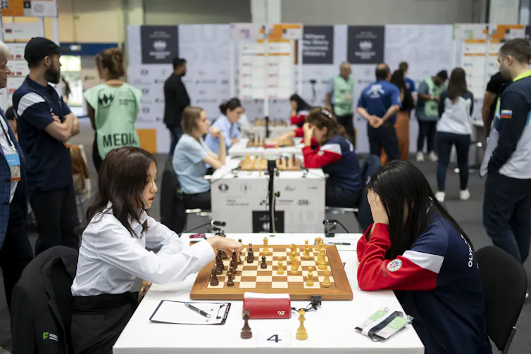 Two young women sit at a table with a chess board between them, and other pairs of players at tables in the background.