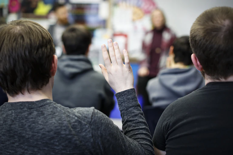 Four boys sit with their backs to the camera, facing two adults standing at the front of a classroom.