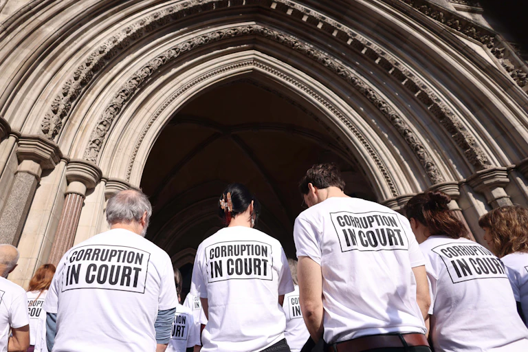 People in white t-shirts stand in front of a stone building with 'corruption in court' on their backs.