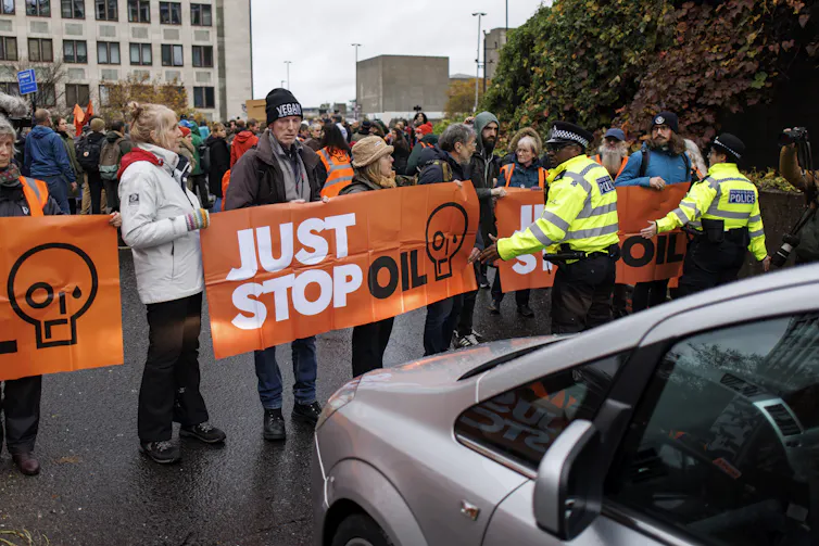 People hold orange Just Stop Oil signs on a road, surrounded by police.