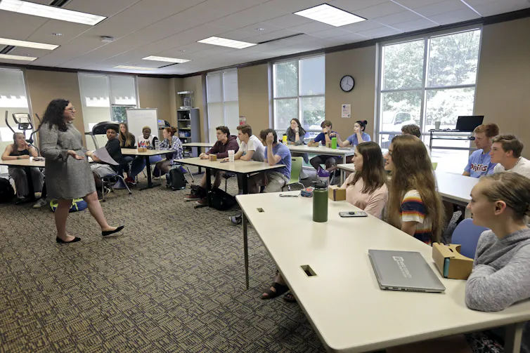 A sunny classroom full of students at long tables, with a female teacher in a dress pacing in front as she talks.