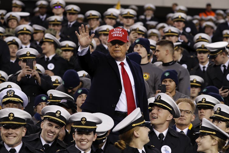 Donald Trump, wearing a dark suit, red tie and cap, standing and waving among uniformed US naval officers.