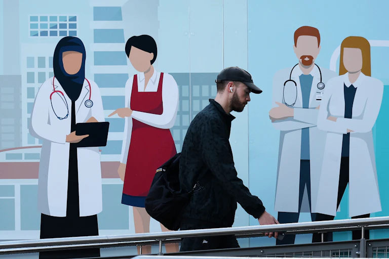 Man in a cap walking up a ramp at an NHS hospital with a mural of healthcare workers on the wall behind him.