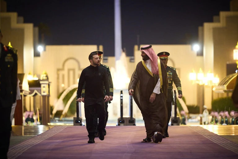 Two men walk on a carpet in front of an illuminated building..