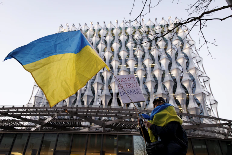A protestor waves a blue and yellow flag and holds a sign that reads "Don't Betray Ukraine".