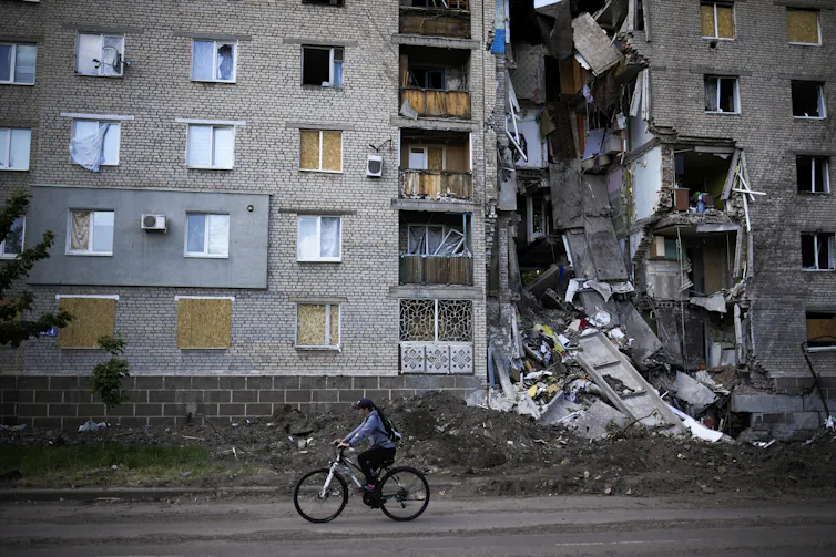 A woman rides a bicycle past a bombed residential building.