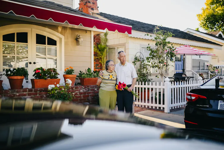 An older man and woman stand outside a suburban house.