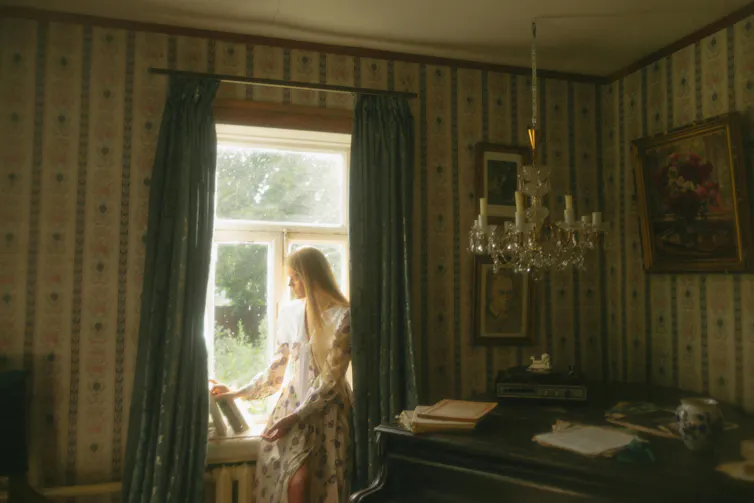 A woman sits by a window, she looks like she is from the 70s.