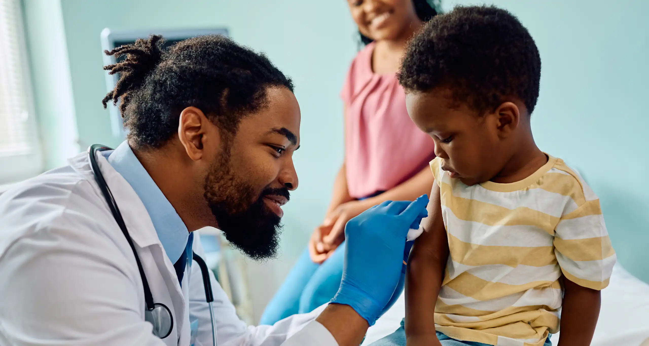 A doctor preparing a child's arm for an injection, with a parent in the background