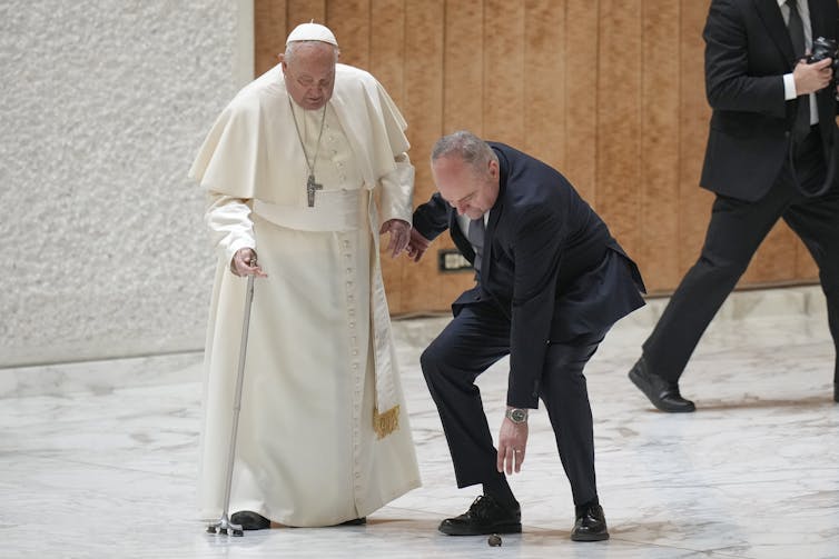 A man in white robes and a large cross with a cane, and a man in a dark suit bending down to pick something up.