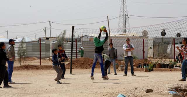 Group of young boys play basketball with refugee camp in background.