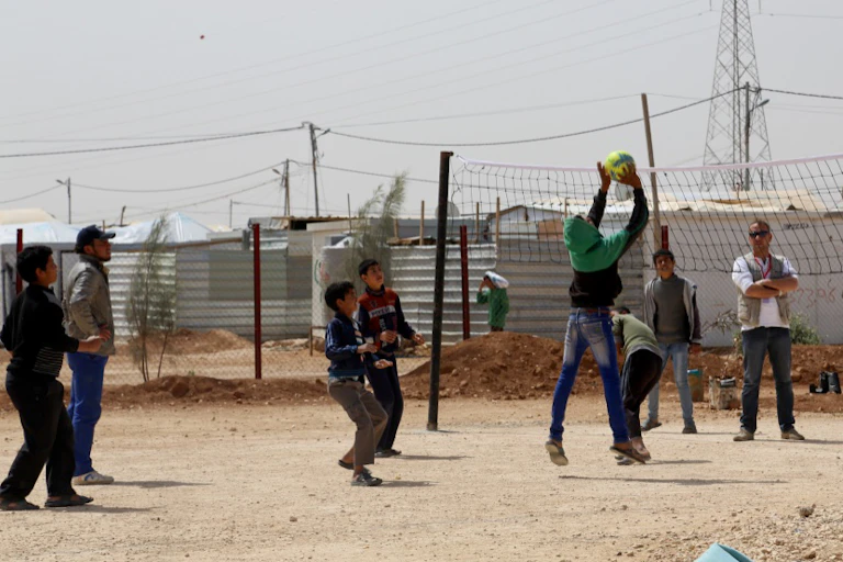 Group of young boys play basketball with refugee camp in background.