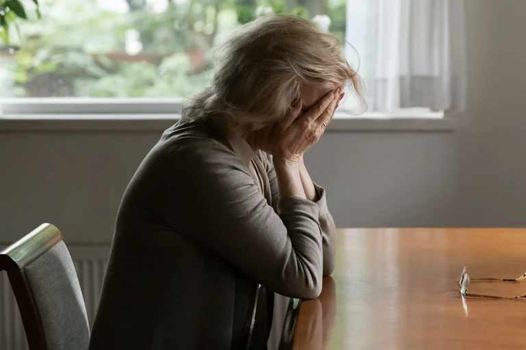 A woman with her head in her hands, seated at a table in her home