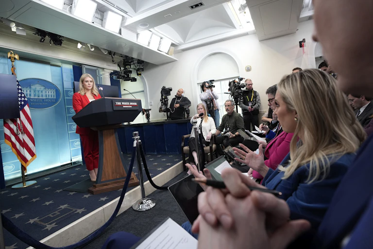 White House press secretary Karoline Leavitt standsw on a stage in the press briefing room talking with jounalists