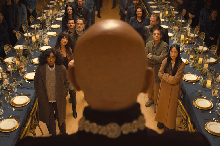 A bald man, his back to us, stands before two long refectory tables filled with attentive young people standing before him.