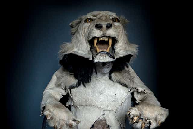 A mannequin wearing a stuffed lion's head as a hat. The lion holds two bags, each with a humanlike head in them.