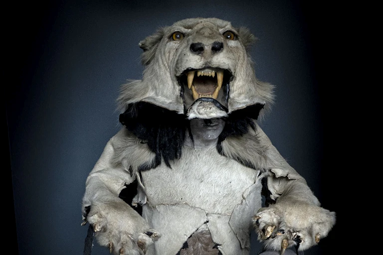 A mannequin wearing a stuffed lion's head as a hat. The lion holds two bags, each with a humanlike head in them.