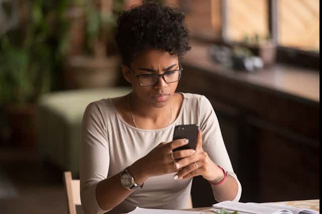 A young black woman who is wearing glasses looks at her smartphone in concern.