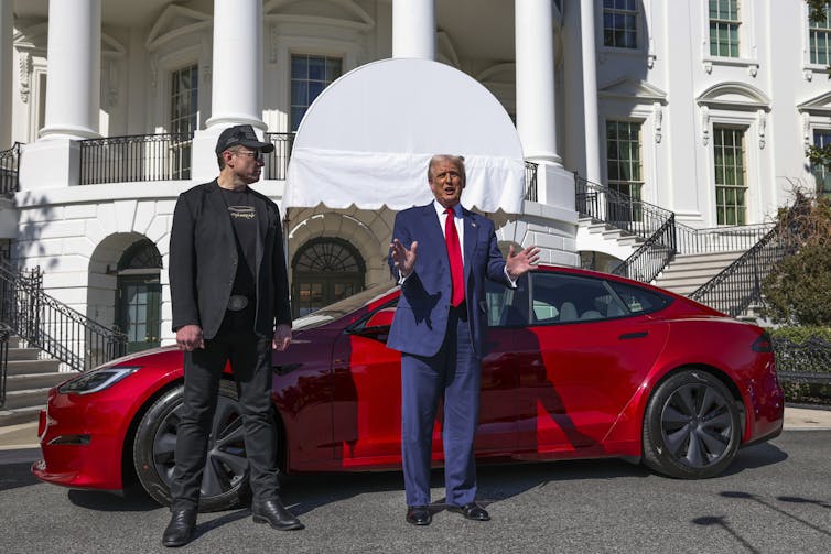 Two men, one wearing black and one wearing a blue suit and red tie, stand in front of a red car outside a large white building.