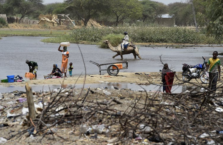A man rides a camel and other people wash clothes at the edge of the lake.