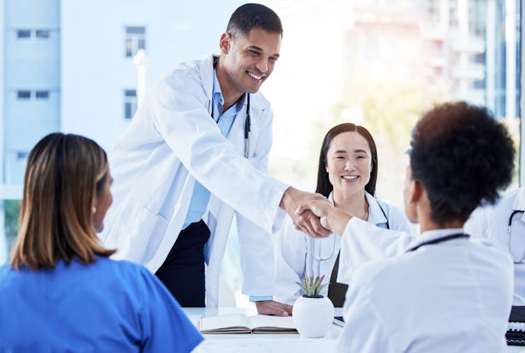 A man in a white coat standing, shaking hands with one of several people seated at a table wearing white coats or scrubs