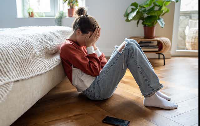A girl sits on the floor leaning against a bed with her face in her palms