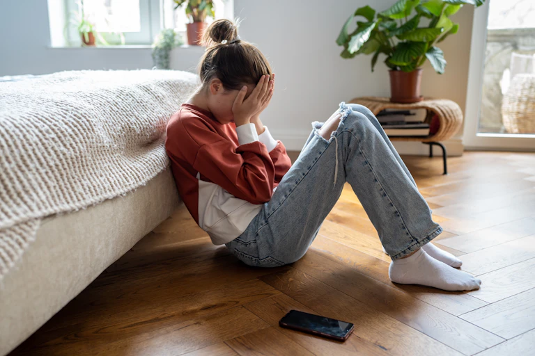 A girl sits on the floor leaning against a bed with her face in her palms