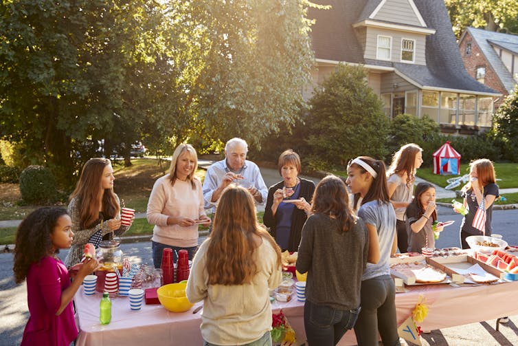 A handful of people of different ages gather around a folding table set on a suburban street.