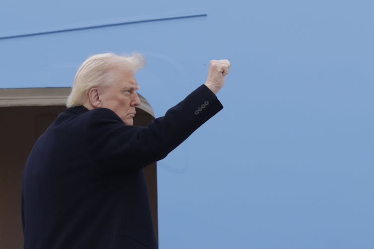 An older man with fluffy white hair gestures with his fist as he walks onto an aircraft.