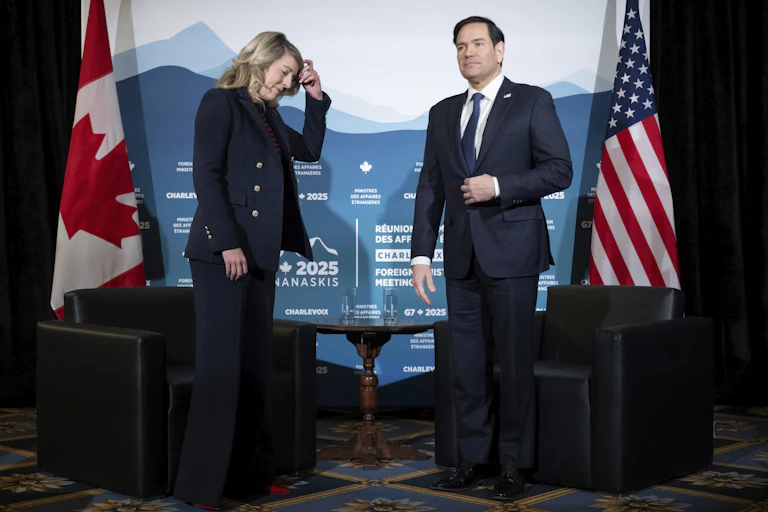 A woman with blonde shoulder-length hair has her hand on her head as she looks down. She's stands next to a dark-haired man. Canadian and U.S. flags are behind them.