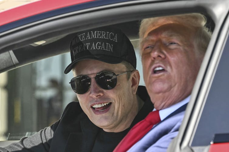 A man in sunglasses and a black ball cap sits inside a red car with an old man with heavy facial bronzer, and orange-tinged hair.