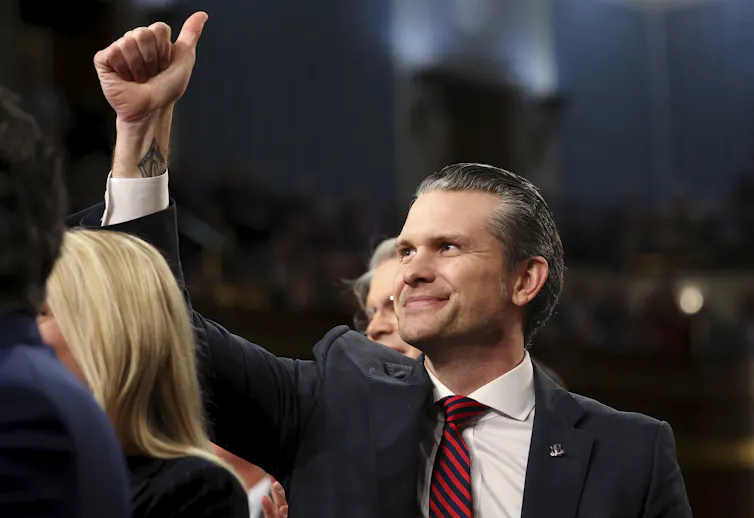 A man with slicked back hair in a suit and tie gives the thumb's up.