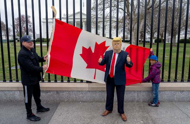 A man and boy hold up a Canadian flag against a wrought-iron fence in front of the White House while another person wears a Trump mask.