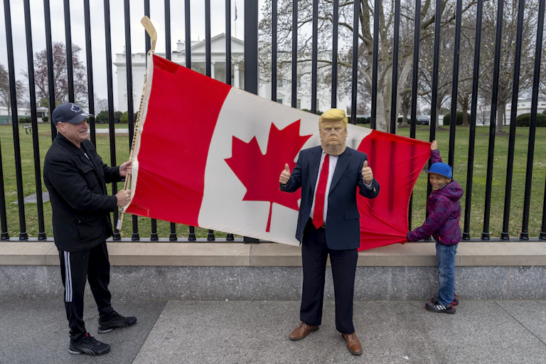 A man and boy hold up a Canadian flag against a wrought-iron fence in front of the White House while another person wears a Trump mask.