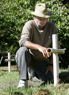 a bearded man in a hat kneels before a cross, greenery behind him