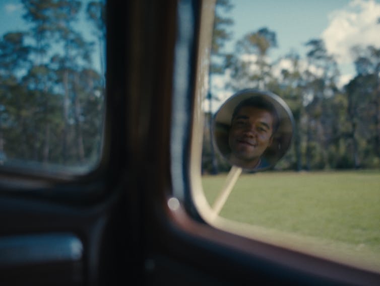 a Black face reflected in a rear-vision mirror, approaching a forest ahead