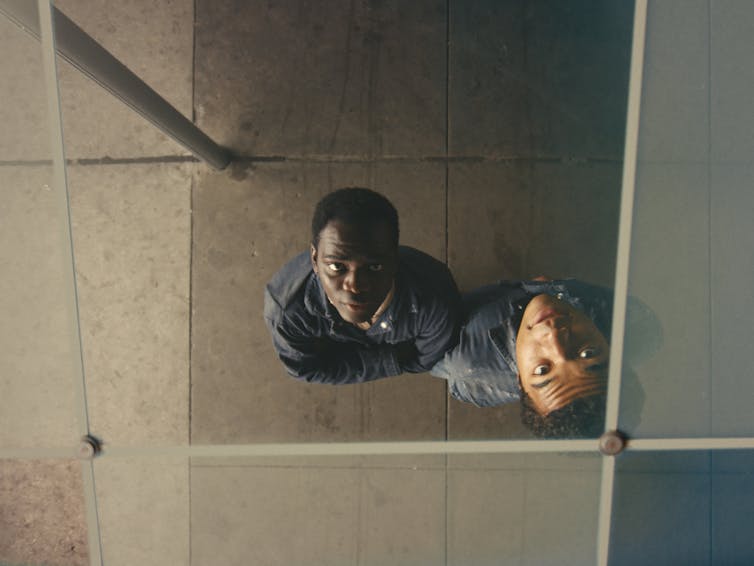 two Black young men, looking up at the camera overhead, from under a glass-tiled roof