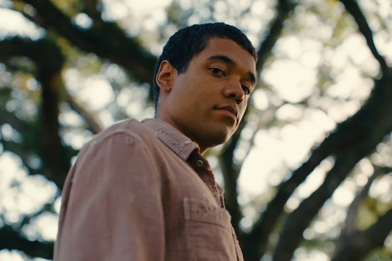 a young man stares at the camera, a tree behind him