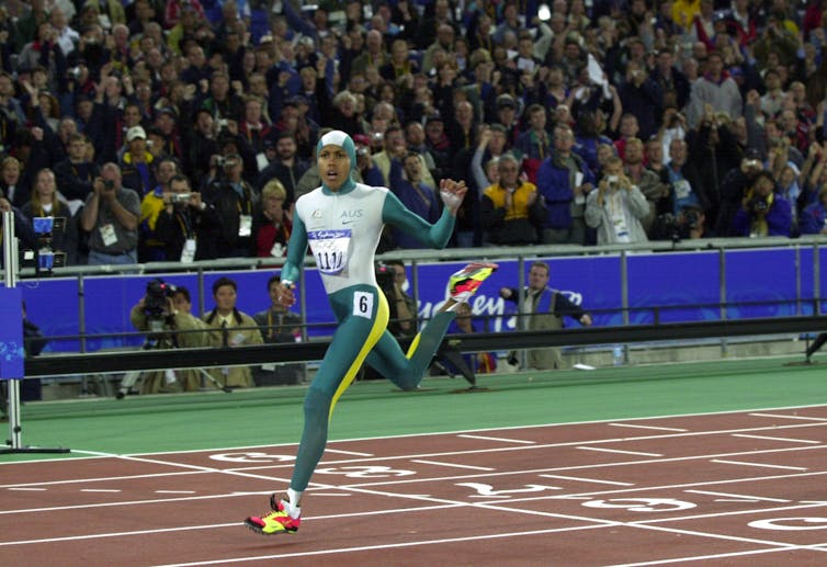 A woman in a full body jumpsuit crosses a sprint finish line in front of a crowd