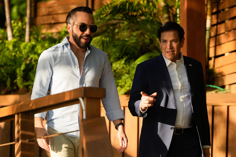 Two men with dark hair, one wearing a dark blazer and the other wearing an open button down shirt, walk together on a leafy path and a wooden building behind them.