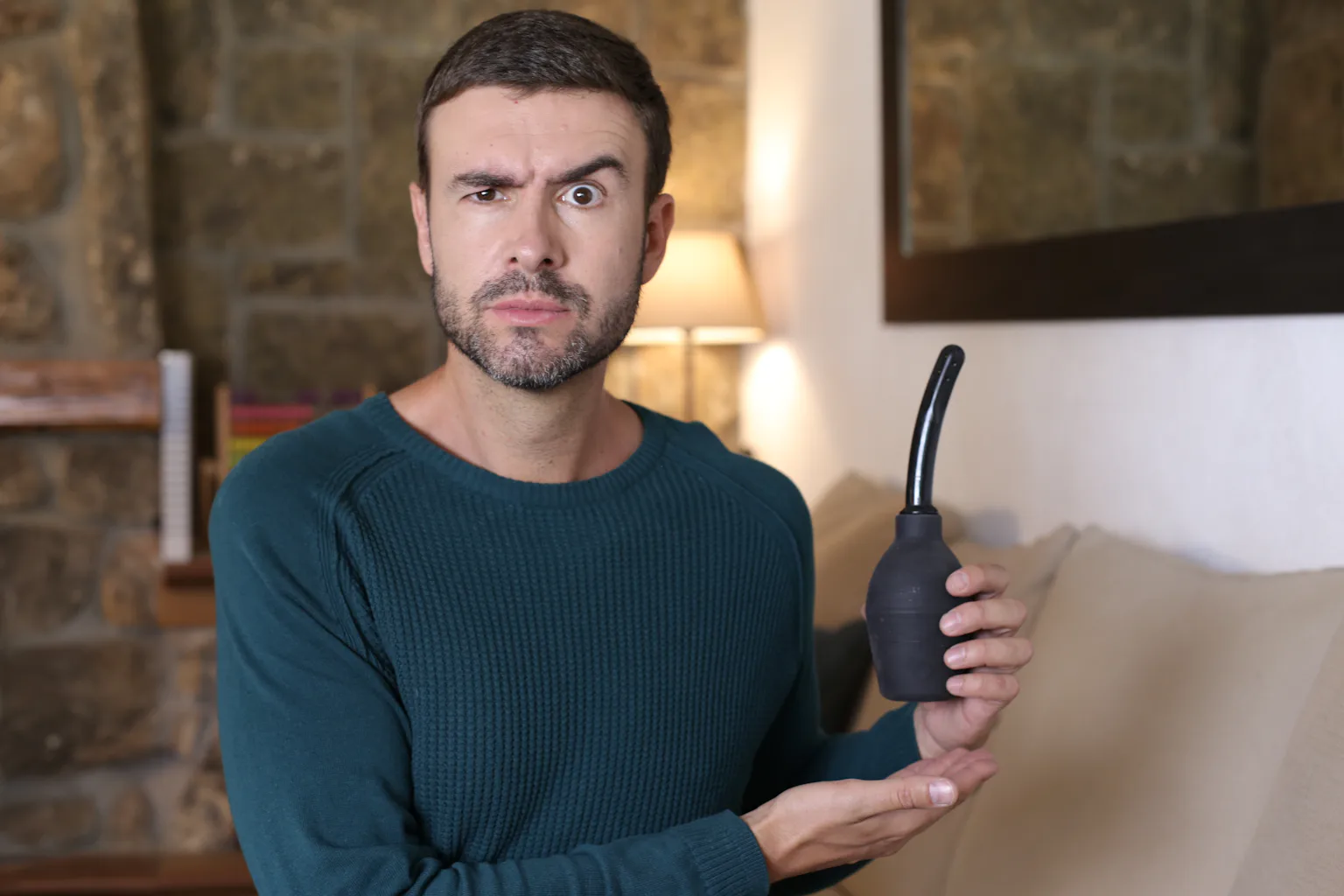 man holding a black rubber enema bulb syringe with a perplexed expression in a home setting