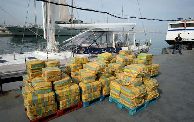 Decenas de fardos de cocaína apilados frente a un velero en el puerto.