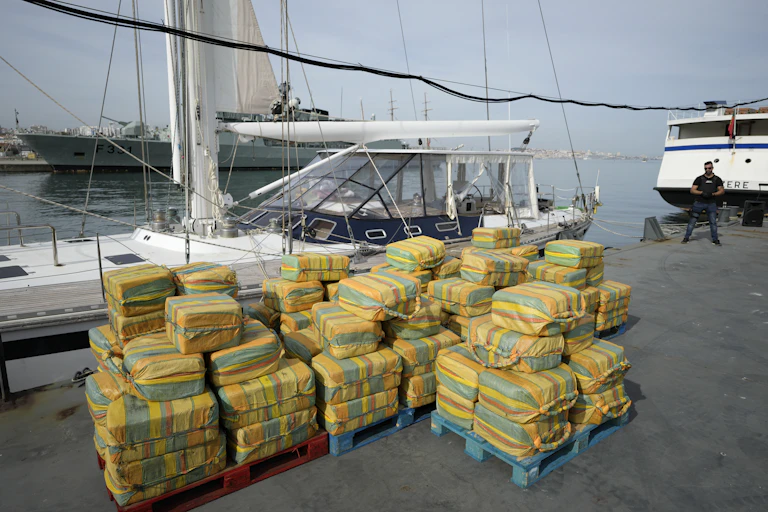 Decenas de fardos de cocaína apilados frente a un velero en el puerto.